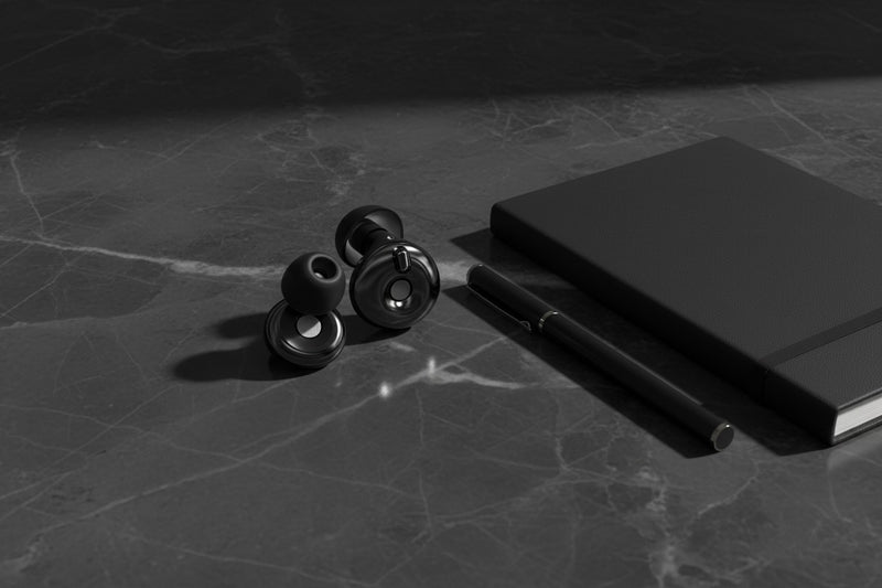 Bouchons d'oreilles QUIET Adapte noirs avec molette ajustable, posés sur une table en marbre près d'un carnet et stylo
