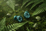 Bouchons d'oreilles QUIET Adapte vert émeraude avec molette ajustable, posés sur de la mousse humide et des fougères.